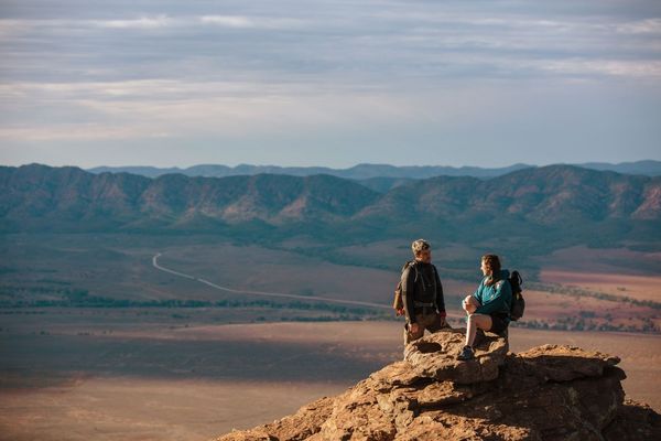 Two walkers resting on a rocky hilltop overlooking the vast red plains and ancient mountain ranges of the Flinders Ranges, South Australia walking holiday